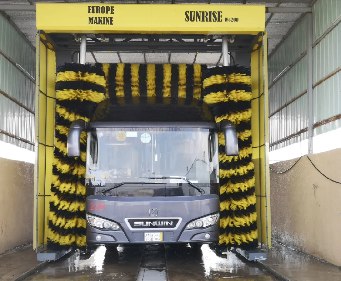 Grey bus inside a yellow wash gantry headed EUROPE MAKINE SUNRISE, with yellow and black brushes closed around its front